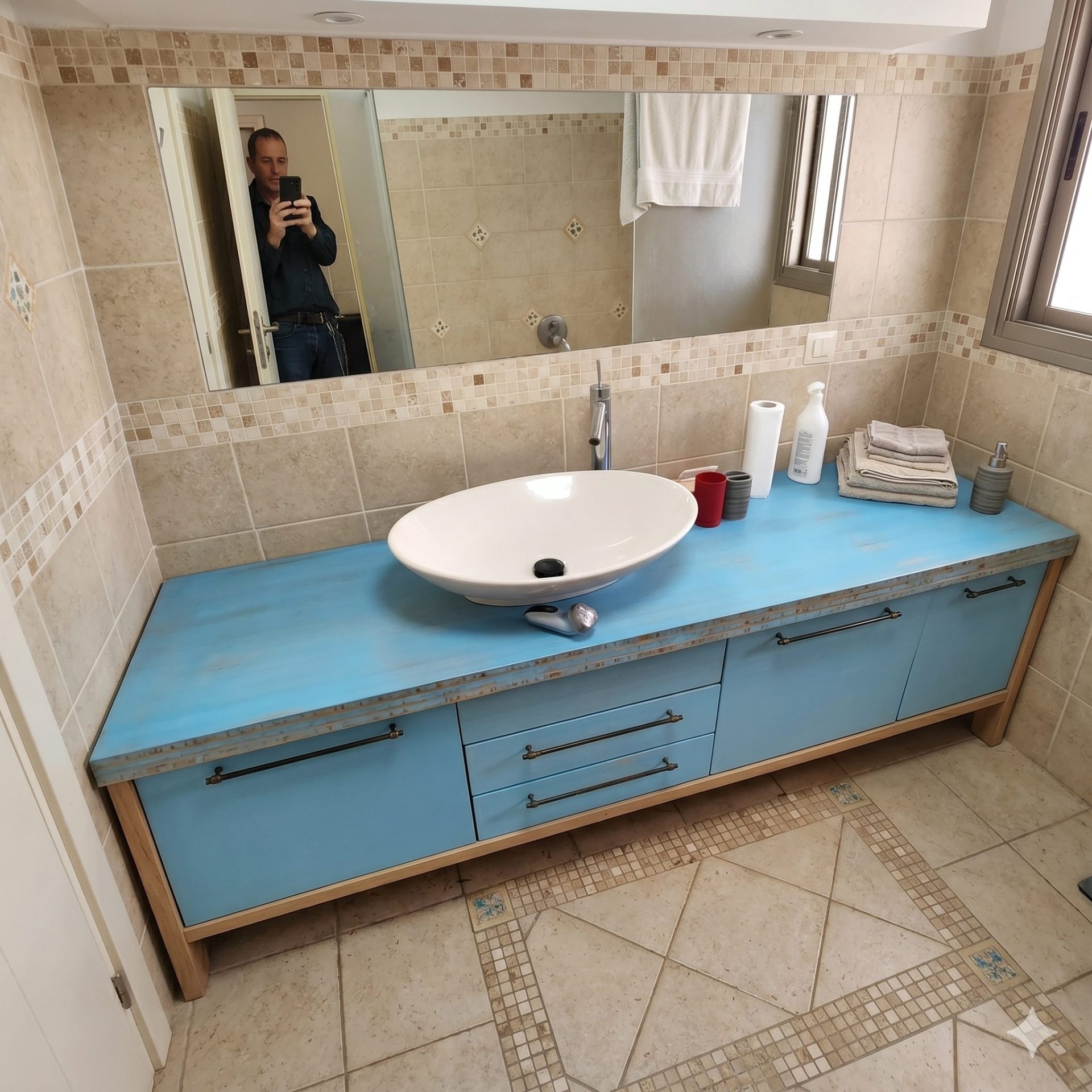 מבט רחב על חדר רחצה הכולל ארון בעיצוב כפרי בצבע תכלת, כיור מונח אובלי לבן, ומראה גדולה שבה משתקף גבר המצלם בטלפון הנייד שלו.