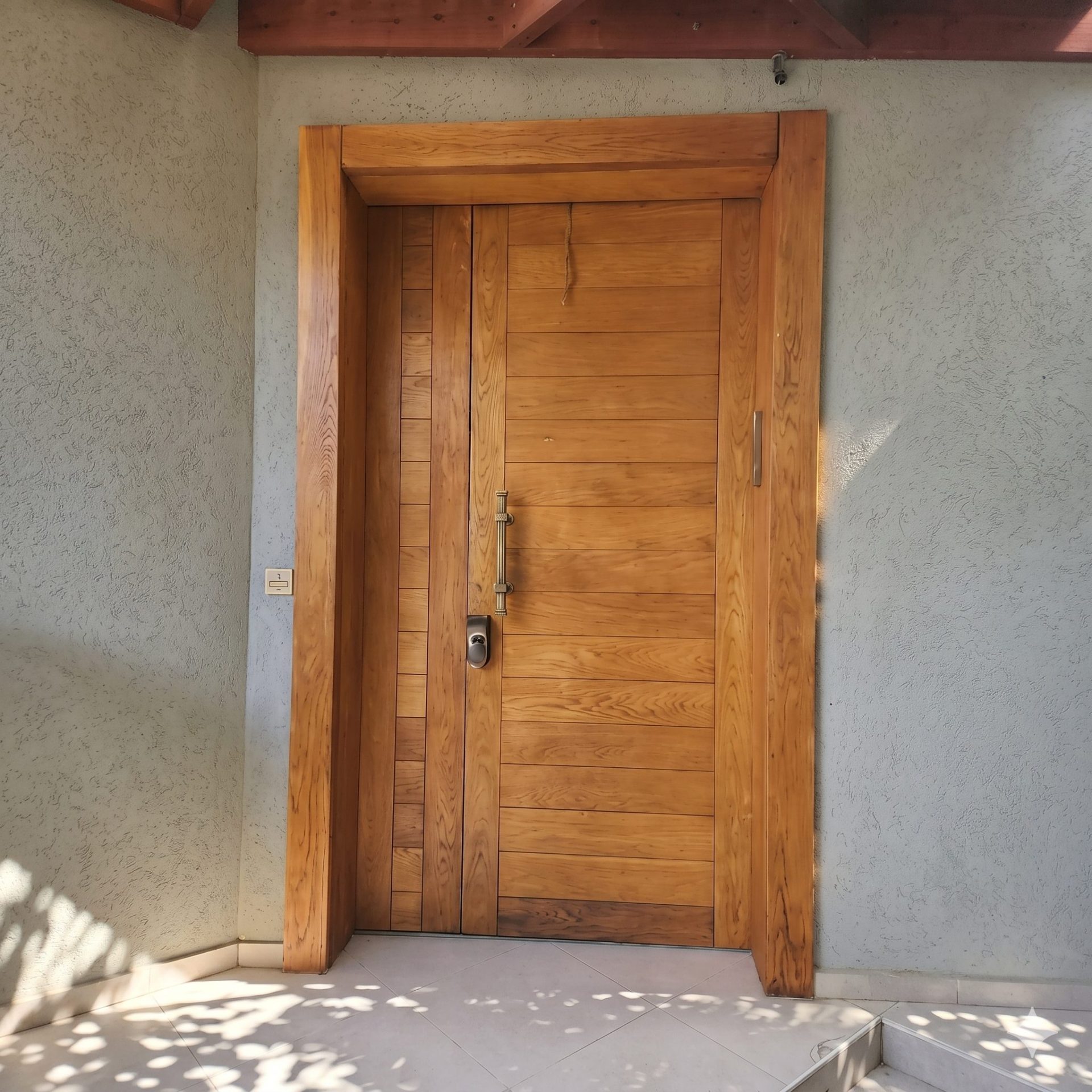 דלת כניסה סגורה מעץ מלא בגוון חם עם ידית אנכית מוארכת ומנעול חכם, מותקנת בתוך משקוף עץ עבה על קיר טיח בהיר.